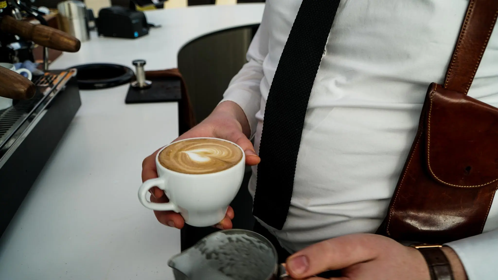Barista in wit overhemd en zwarte stropdas maakt latte art in een kopje cappuccino aan de bar.