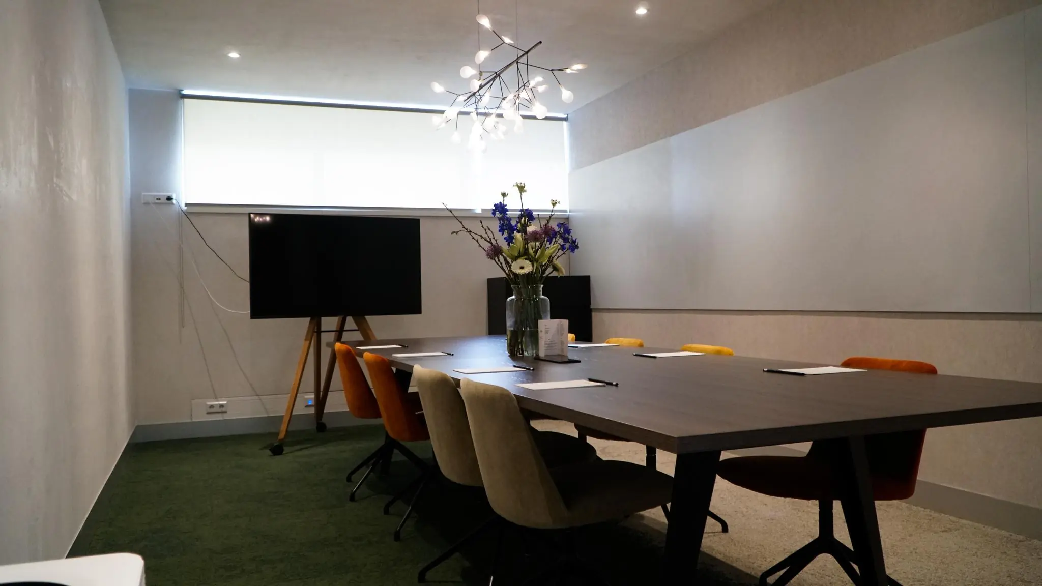 Moderne vergaderruimte in Bisonspoor met lange tafel, gekleurde stoelen, bloemenvaas en flatscreen op standaard.