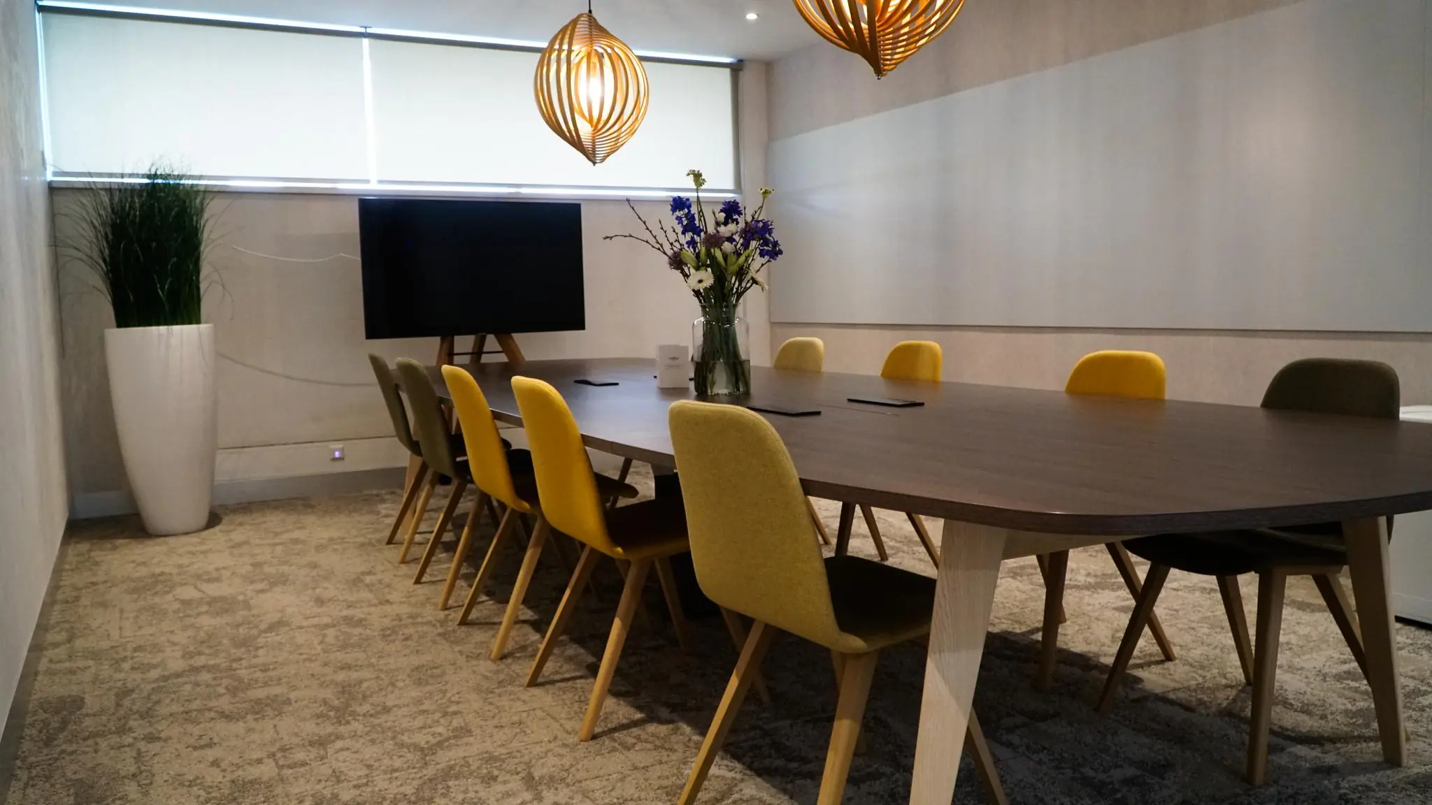 Vergaderruimte Bisonspoor met lange houten tafel, gele en groene stoelen, bloemenvaas, televisie en moderne verlichting.