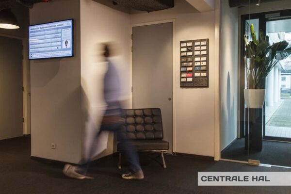Centrale hal van het Business Centrum Huizen met bewegende persoon, digitale informatiebord en zitbank.