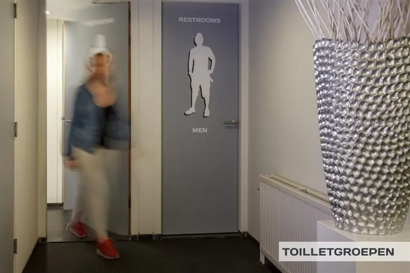 Gang met deuropeningen naar herentoilet en een persoon die voorbijloopt in een kantooromgeving aan de Huizermaatweg.