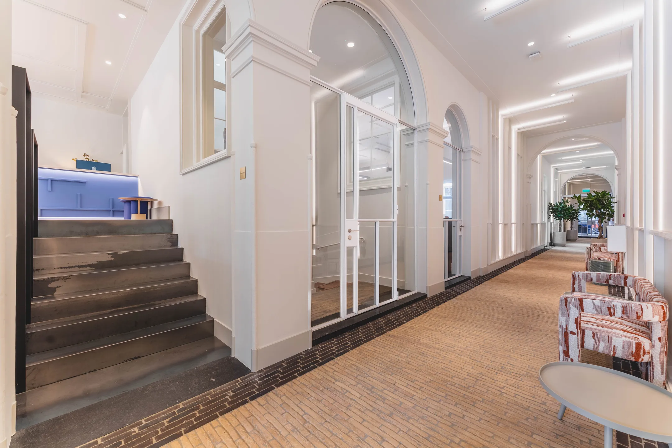 Een moderne, lichte gang met een blauwe balie, stoelen en groene planten.