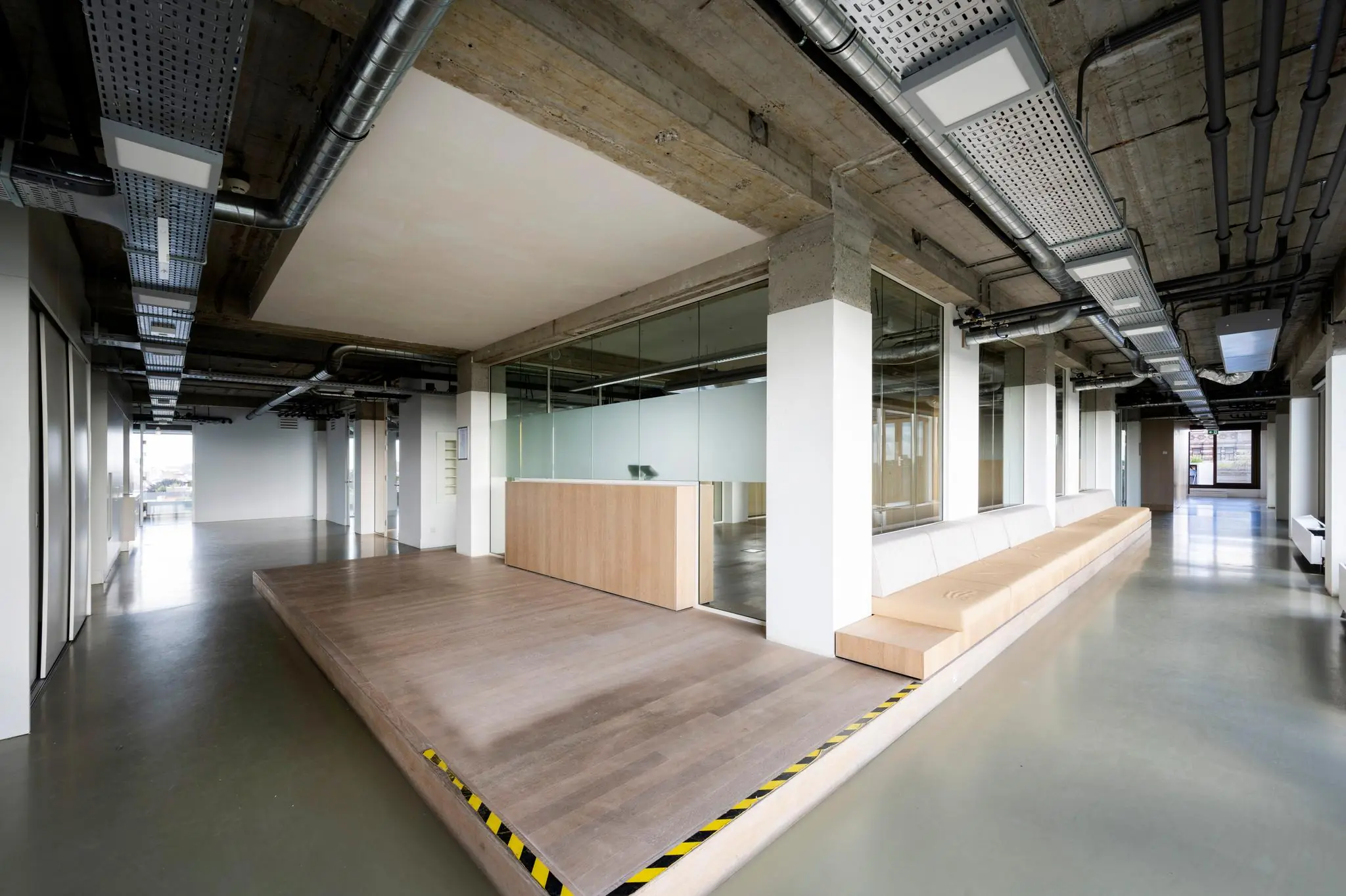 Moderne kantoorruimte aan de Vijzelstraat met een verhoogd houten platform, glazen wanden en een lange zitbank.