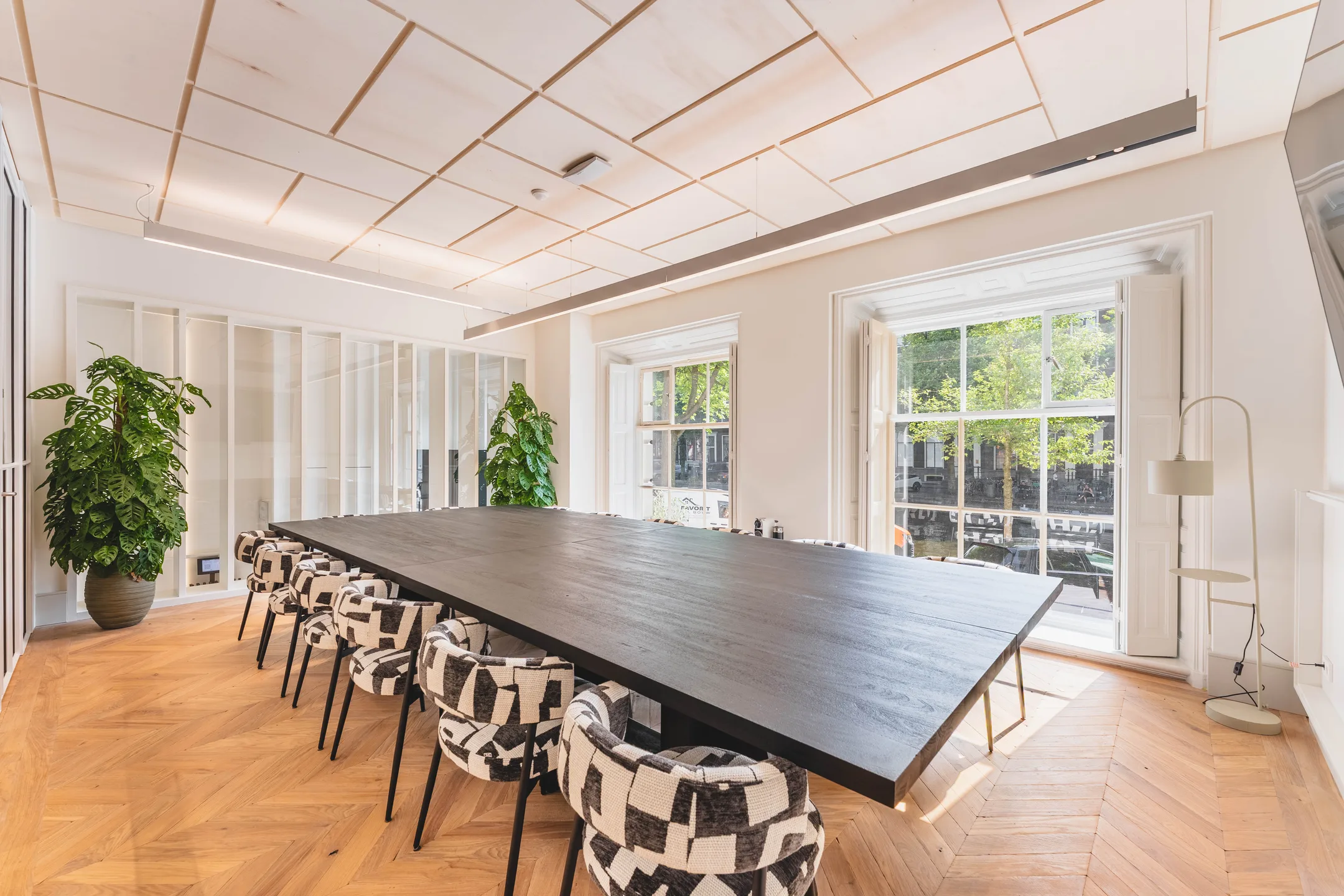 Lichte vergaderruimte met een grote houten tafel, designstoelen en twee grote ramen die uitzicht bieden op groen buiten.