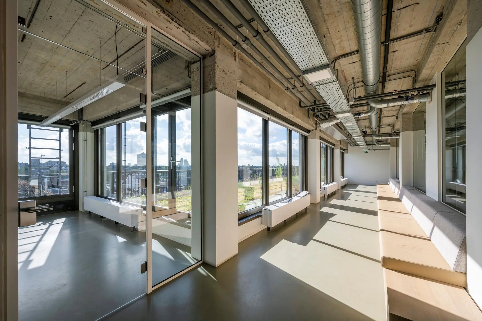 Modern kantoorinterieur aan de Vijzelstraat met grote ramen, een betonnen plafond en ingebouwde zitbanken langs de ramen.