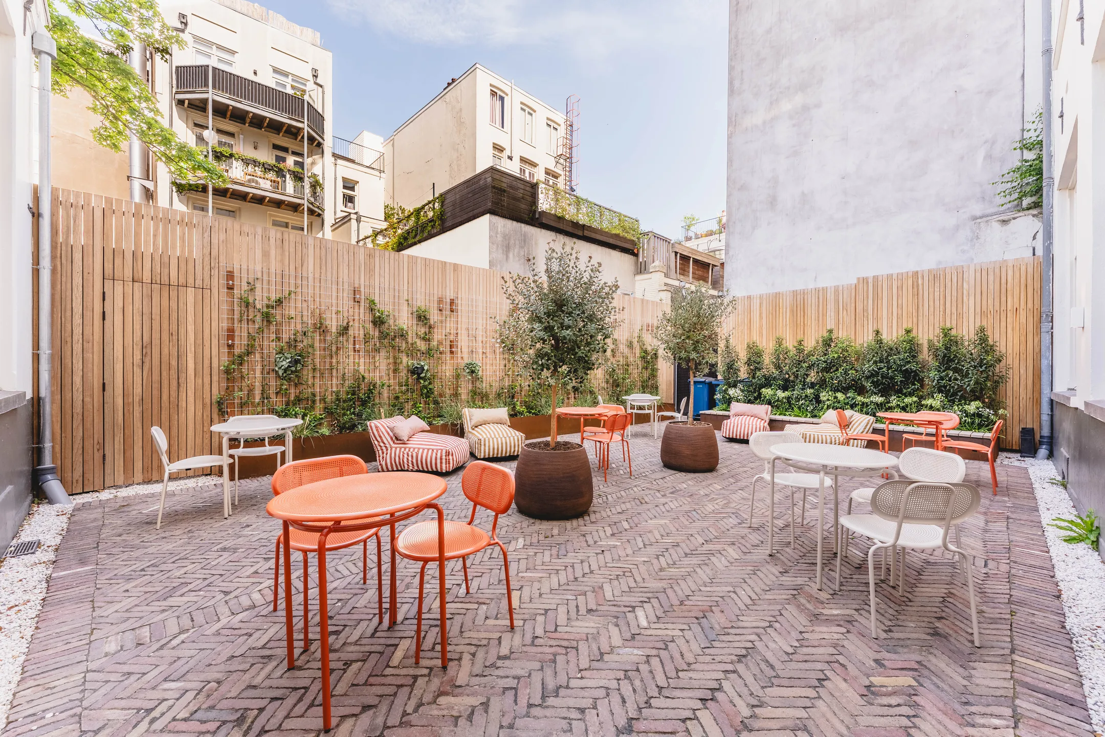 Een zonnige binnenplaats met kleurrijke stoelen, tafels en planten tegen een houten omheining.