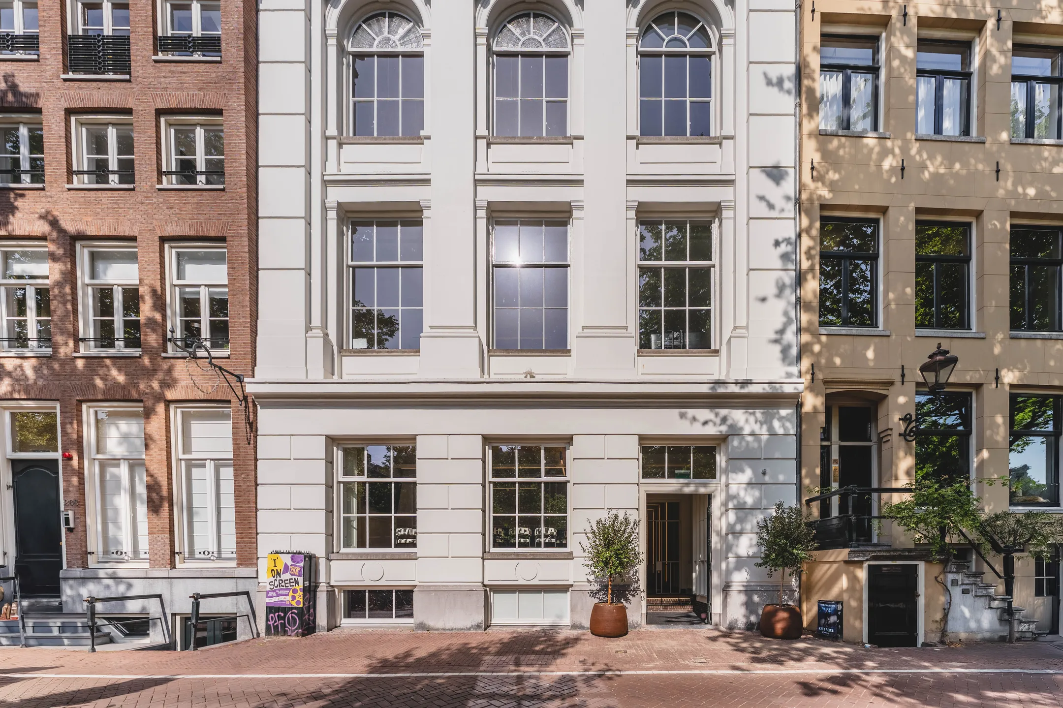 Een historische gevel van een pand tussen twee andere gebouwen met schaduwen van bomen op de gevels.