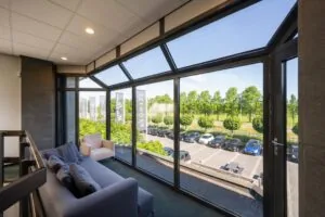 Zithoek met bank en stoel bij grote glazen ramen met uitzicht op parkeerplaats en groene omgeving bij Stammerhove.