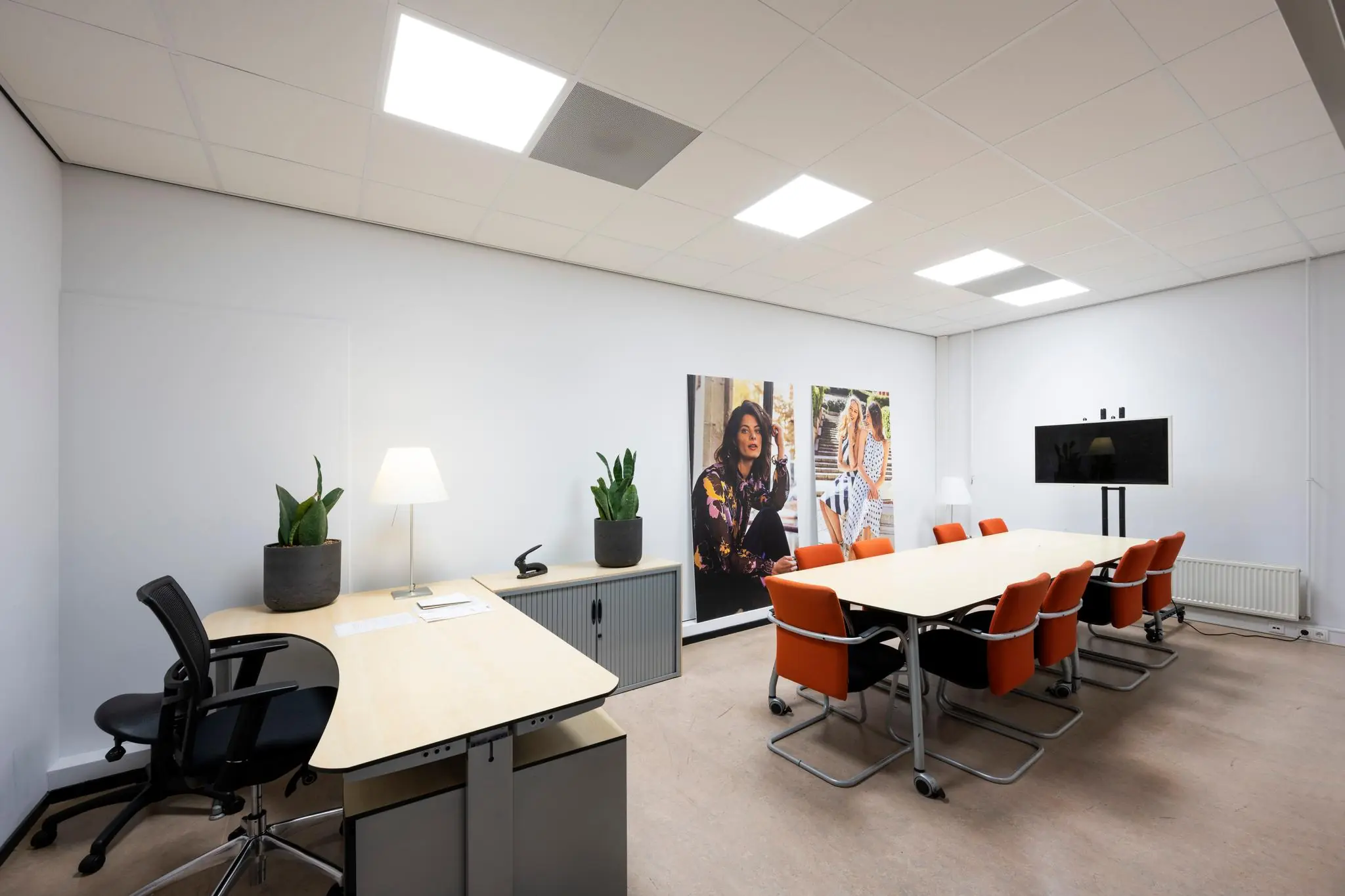 Moderne vergaderruimte met lange tafel, oranje stoelen, bureau, muurdecoratie en groot videoscherm.
