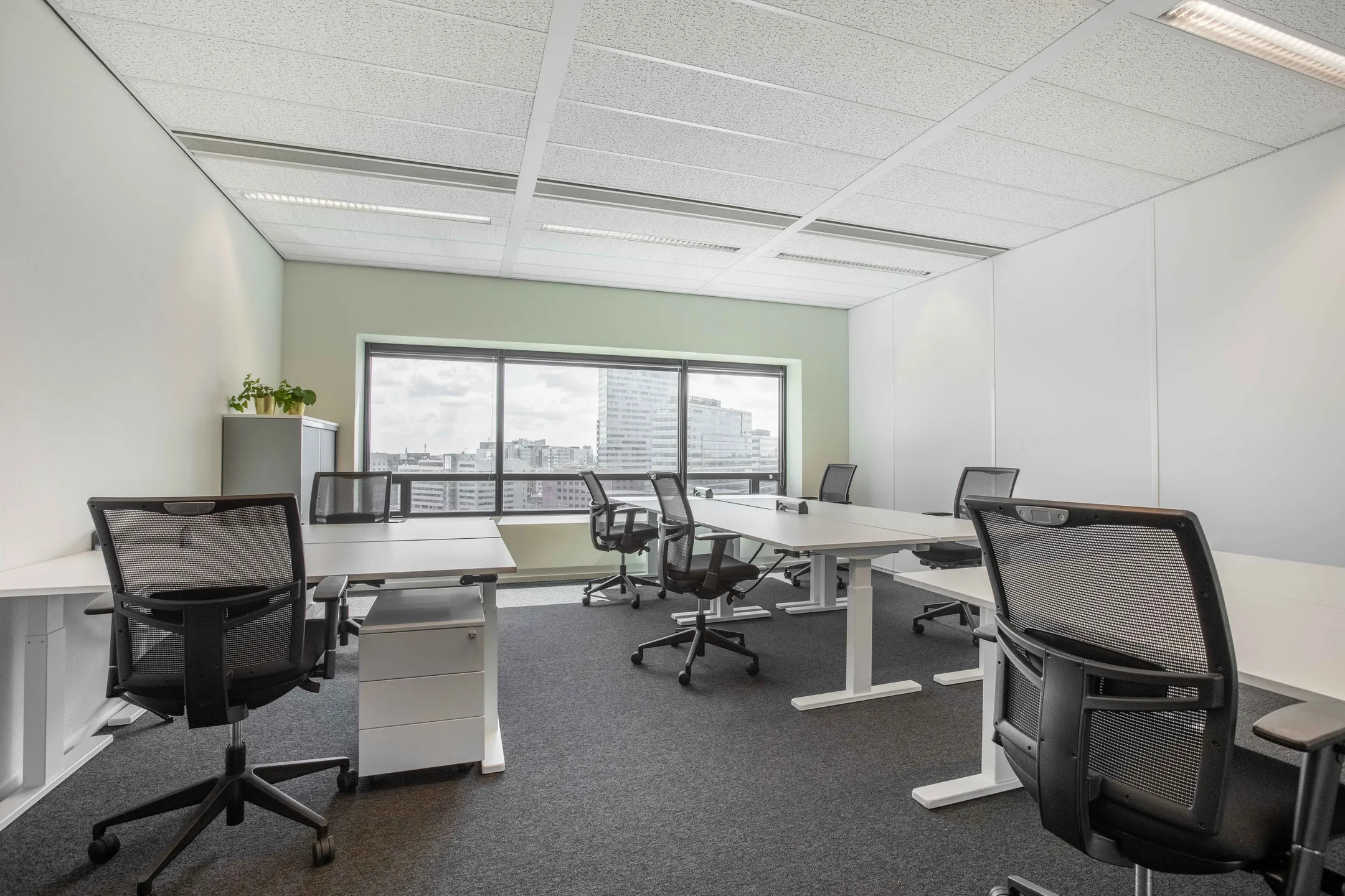 Een modern en licht kantoor met meerdere bureaus en stoelen, en een raam met uitzicht op de stad.