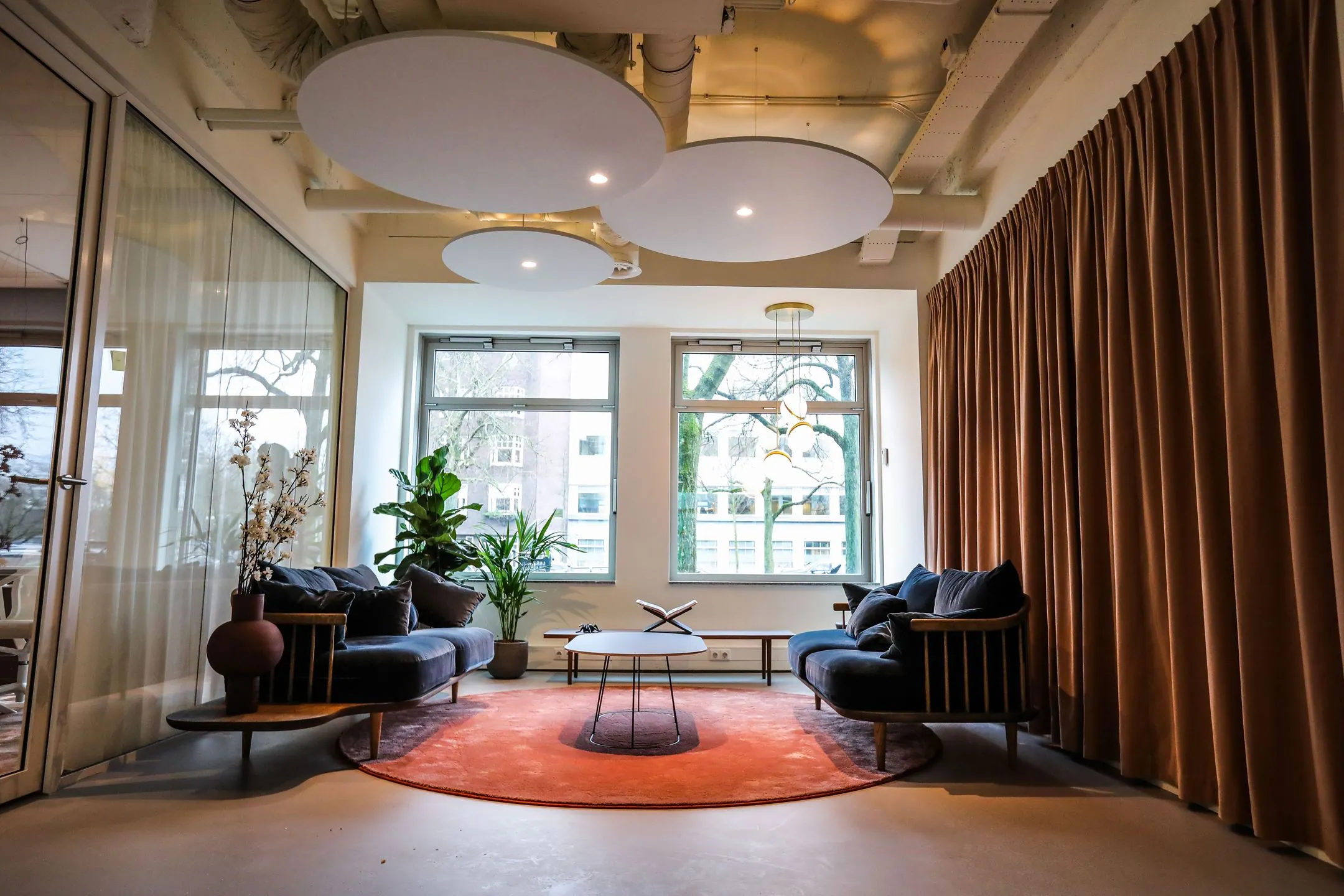 Stijlvolle wachtruimte met twee donkerblauwe banken, een ronde oranje vloerkleed en grote ramen met uitzicht op bomen.