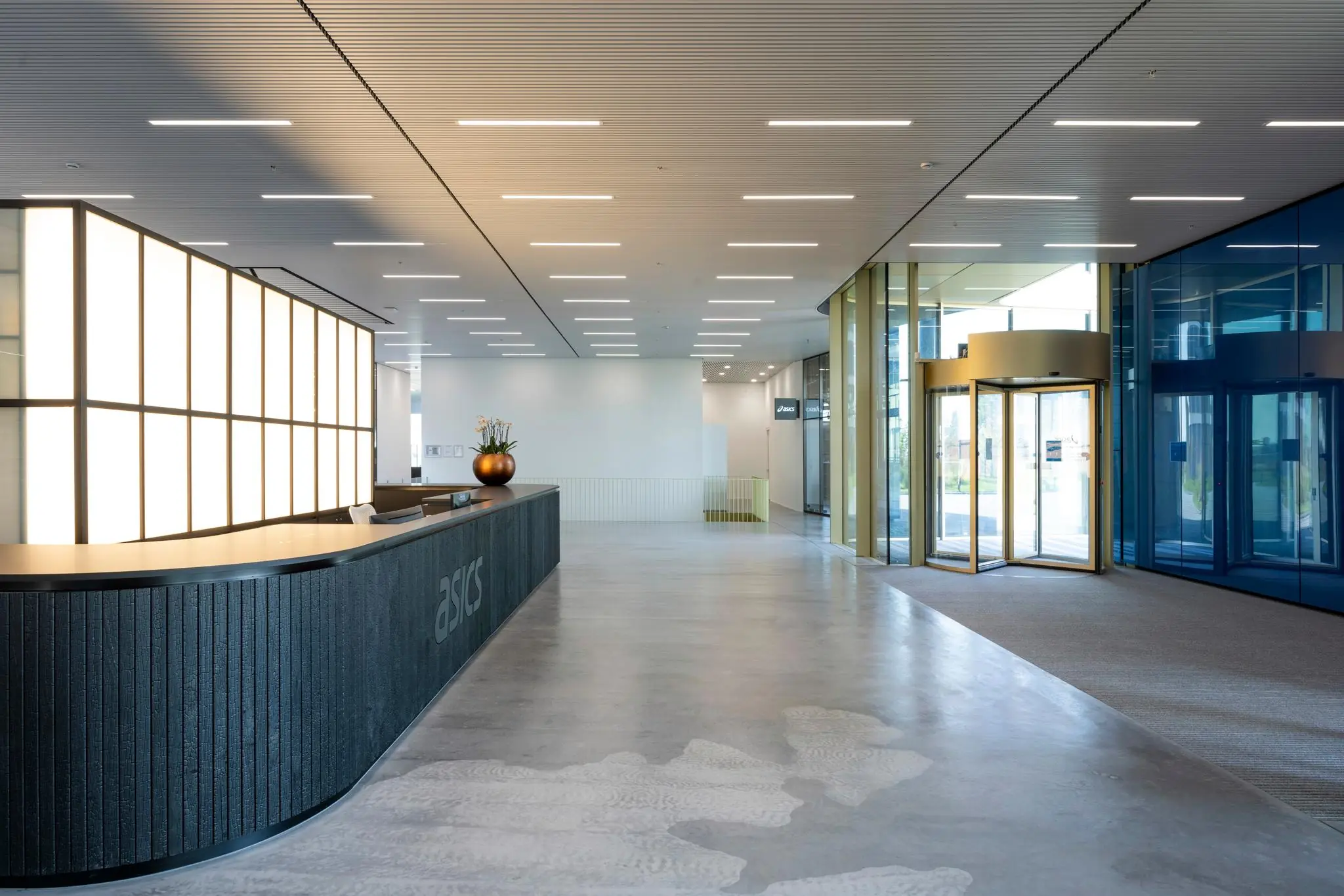 Moderne ontvangsthal van het ASICS-kantoor aan Taurusavenue met een ronde balie, glazen draaideur en strakke verlichting.