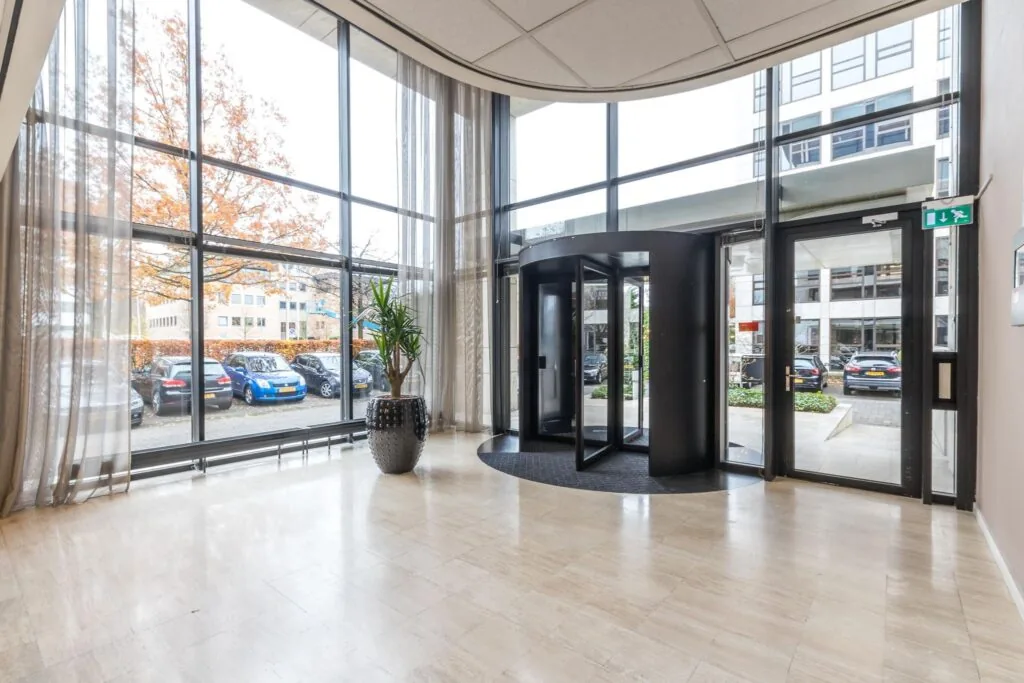 Entree van een modern kantoorgebouw met een draaideur, grote ramen en uitzicht op geparkeerde auto's.