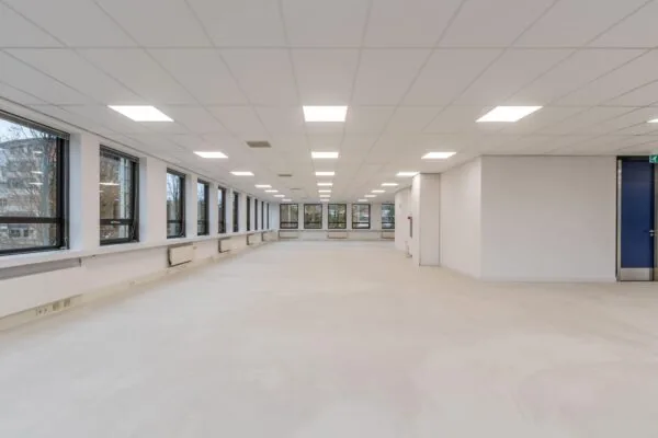 Lichte, lege kantoorruimte met systeemplafond, grote ramen en een betonvloer.
