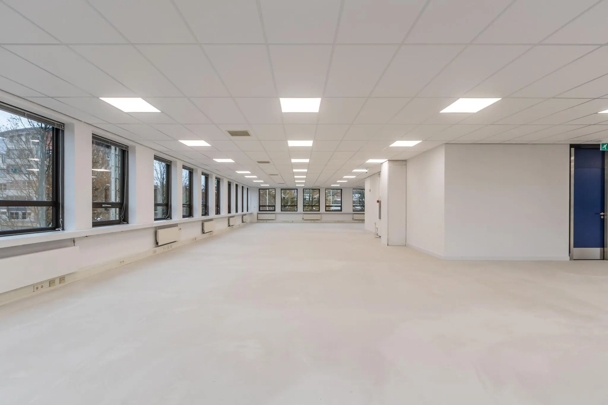 Lichte, lege kantoorruimte met systeemplafond, grote ramen en een betonvloer.