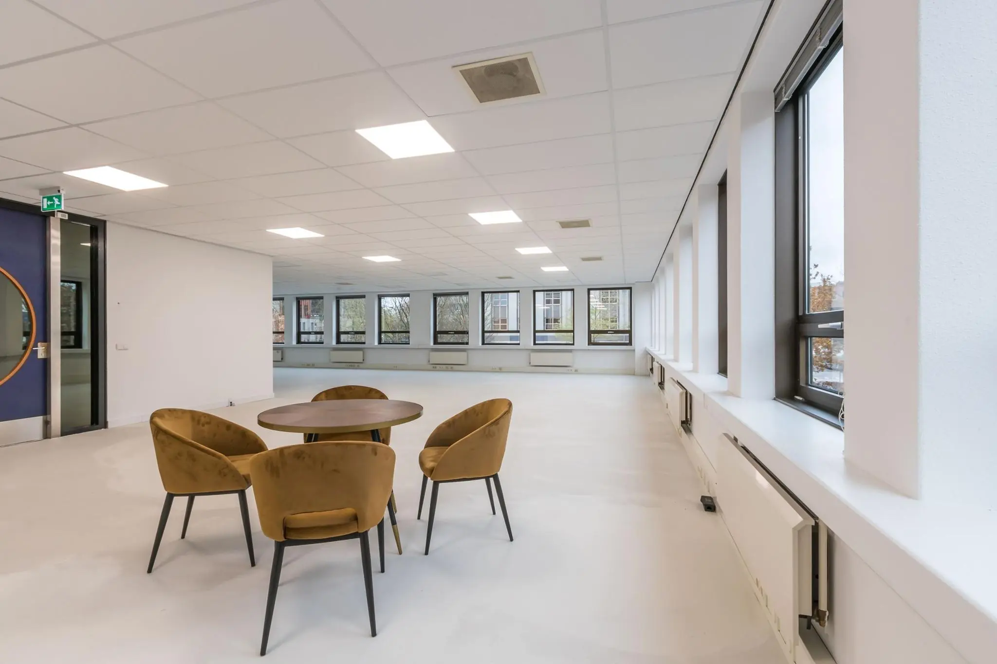 Moderne, lichte kantoorruimte met ronde tafel en vier fluwelen stoelen bij een rij grote ramen.