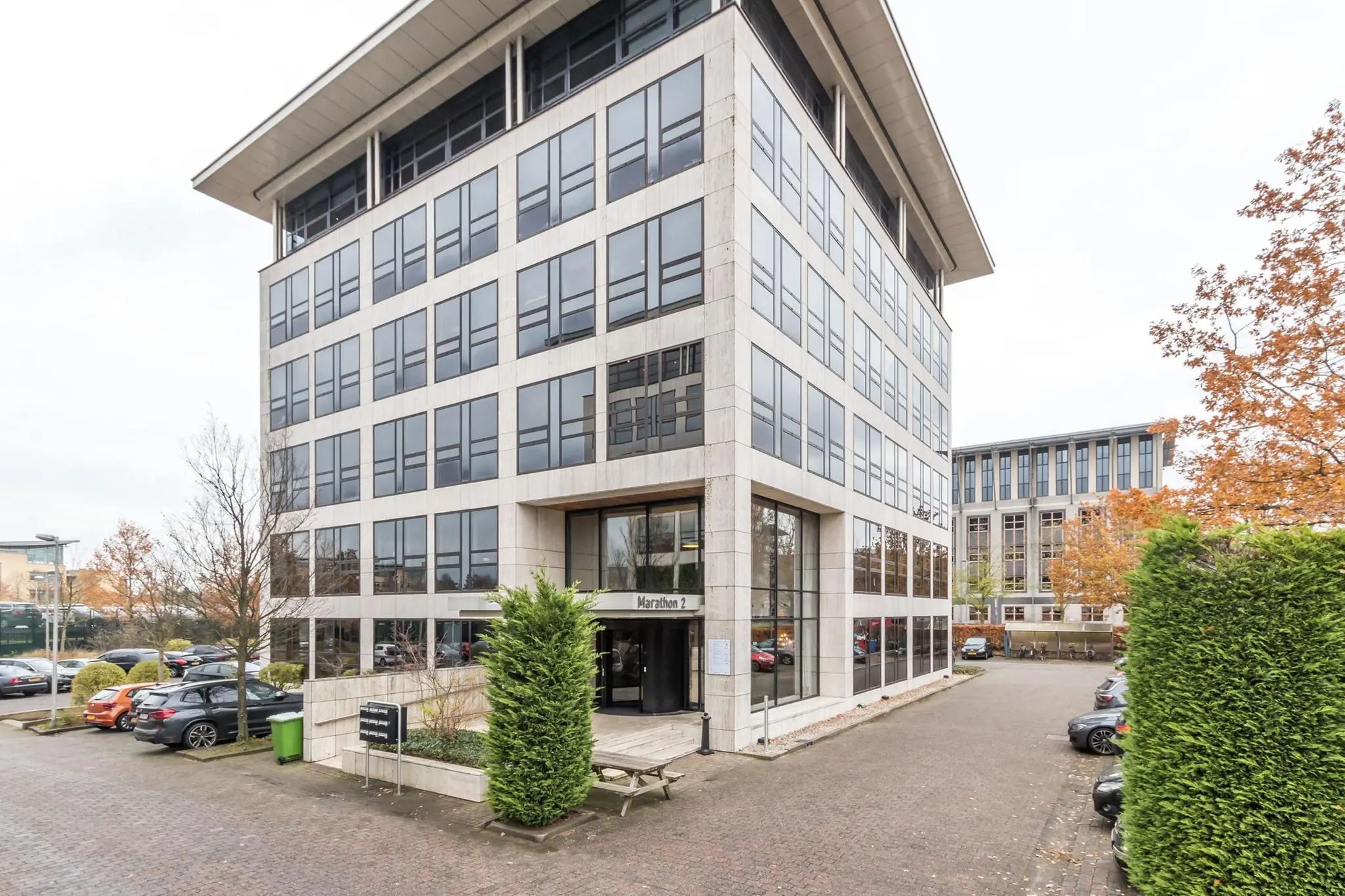 Vooraanzicht van kantoorgebouw Marathon 2 met parkeerplaatsen en herfstbomen.