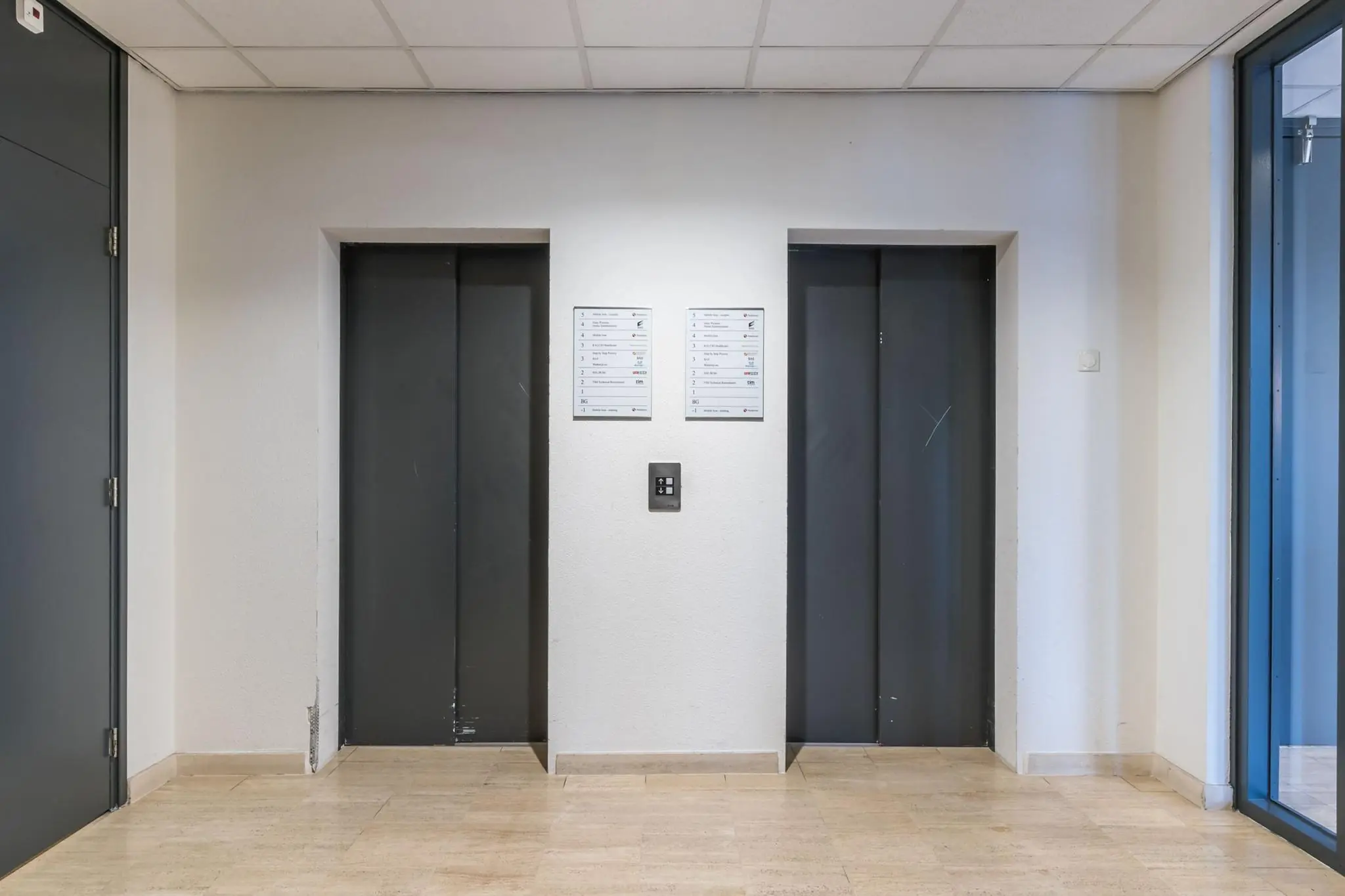 Twee gesloten, donkere liftdeuren in een strak afgewerkte gang met beige tegelvloer en witte muren.