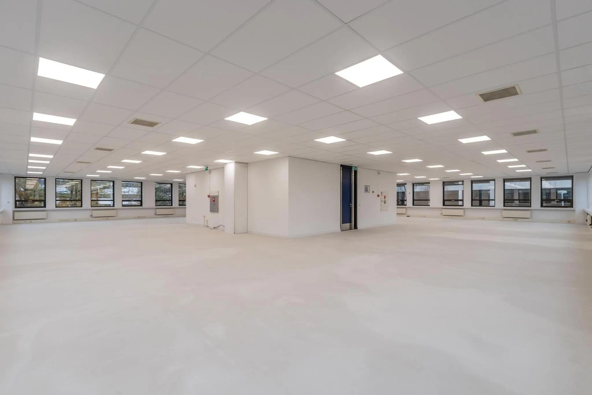 Lichte, lege kantoorruimte met systeemplafond en grote ramen aan twee zijden.