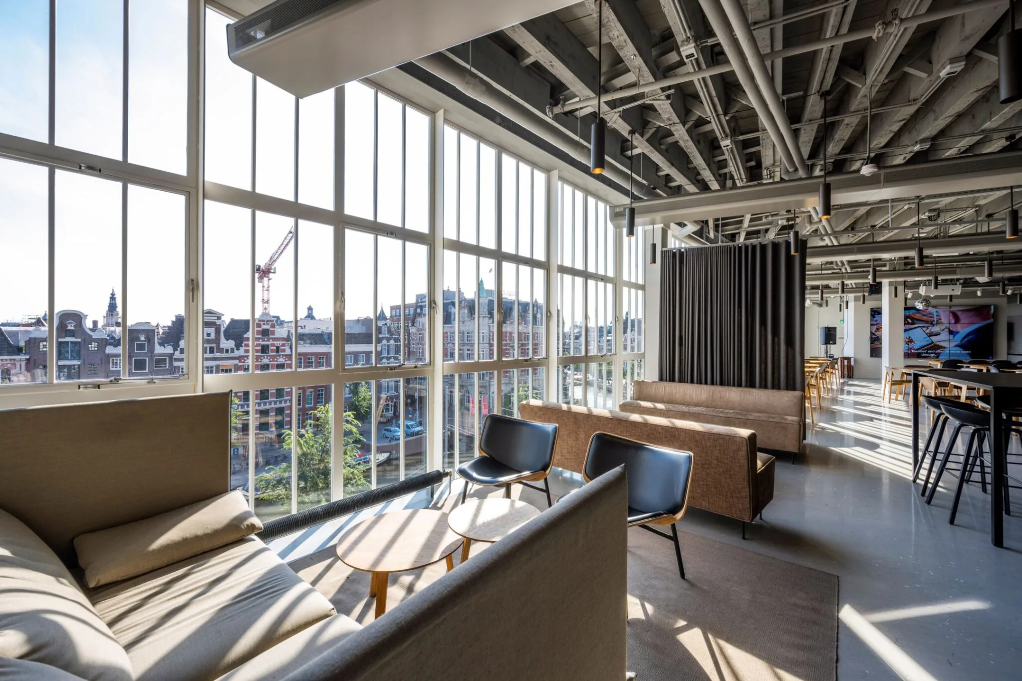 Lichte en moderne loungeruimte met uitzicht op Amsterdamse grachtenpanden door hoge ramen.