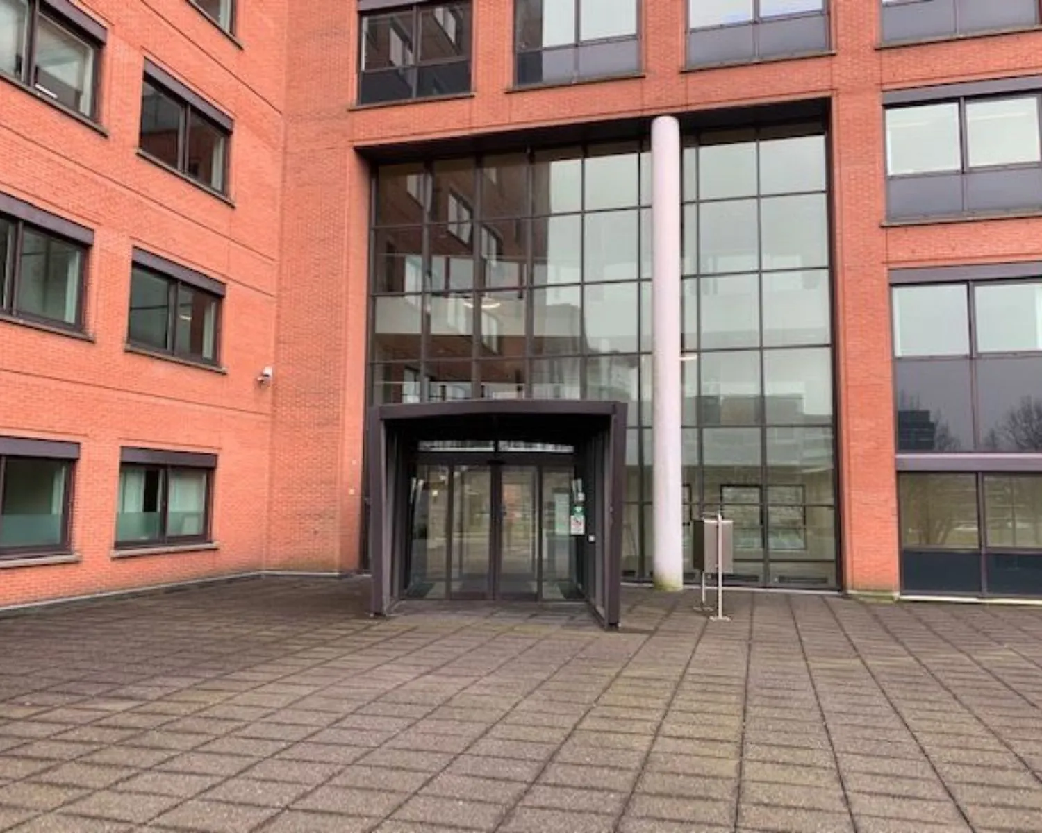 Glazen entree van een modern rood bakstenen kantoorgebouw met draaideur en tegelplein.