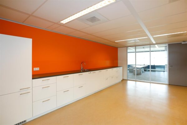 Moderne kantoorkeuken met witte kasten, een feloranje achterwand en uitzicht op een vergaderruimte achter glas.