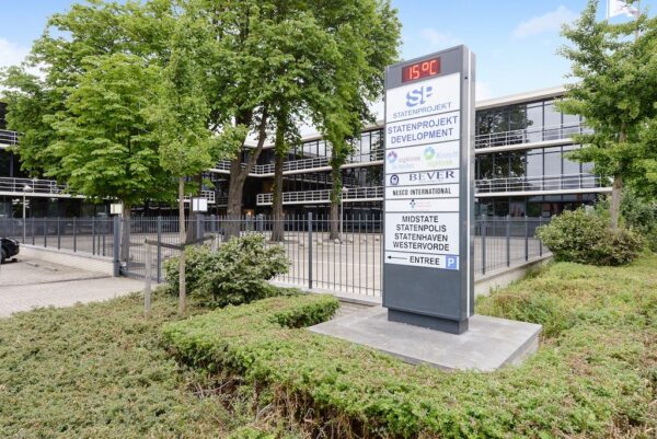 Informatiebord met bedrijfsnamen voor een kantoorpand aan de Rijksstraatweg, omringd door bomen en hekwerk.
