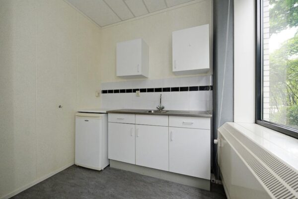 Kleine kitchenette met witte keukenkastjes, een compacte koelkast en een raam dat uitkijkt op groen.