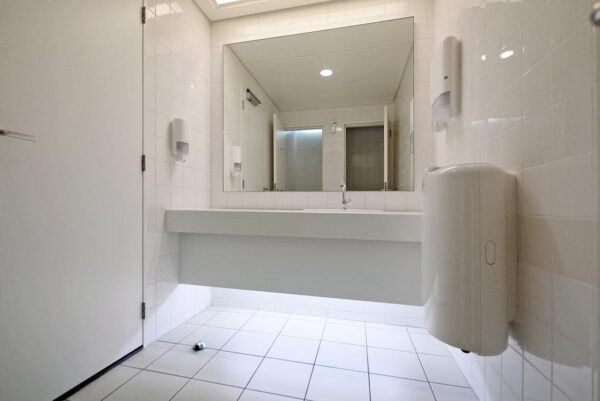 Een moderne, wit betegelde openbare toiletruimte met een grote spiegel, wasbak en handdroger.