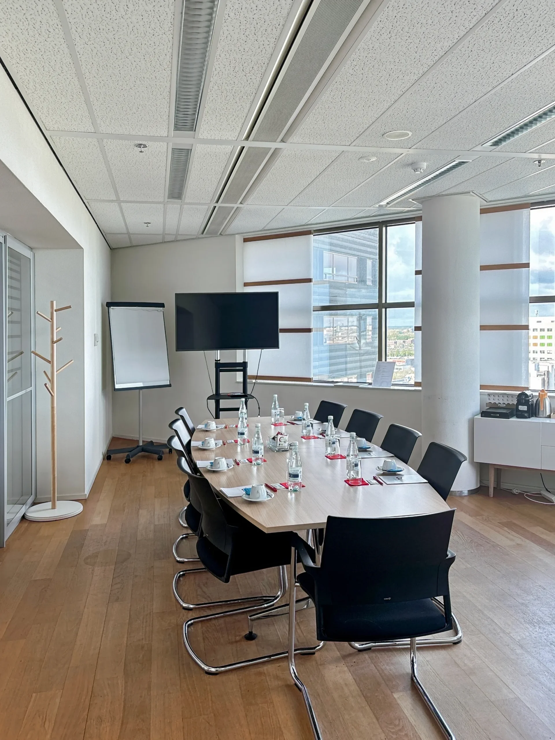 Vergaderruimte met een ovale tafel, stoelen, flesjes water en een flip-over bij het raam.