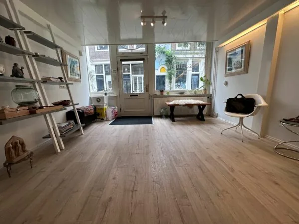 Lichte en modern ingerichte wachtruimte met houten vloer, boekenplanken en grote ramen met zicht op de straat.