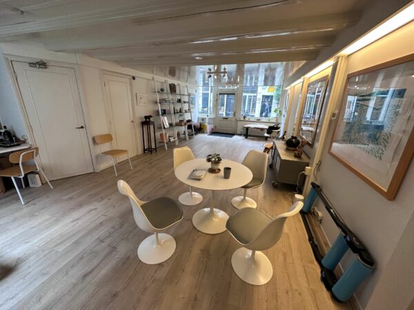 Moderne, licht ingerichte ruimte met een ronde tafel, designstoelen en een wand met kunst en grote ramen aan de straatzijde.