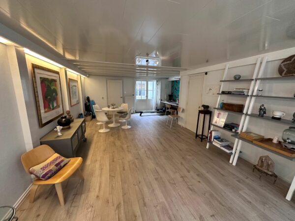 Moderne, langgerekte woonkamer met houten vloer, witte meubels en een open boekenkast tegen de muur.