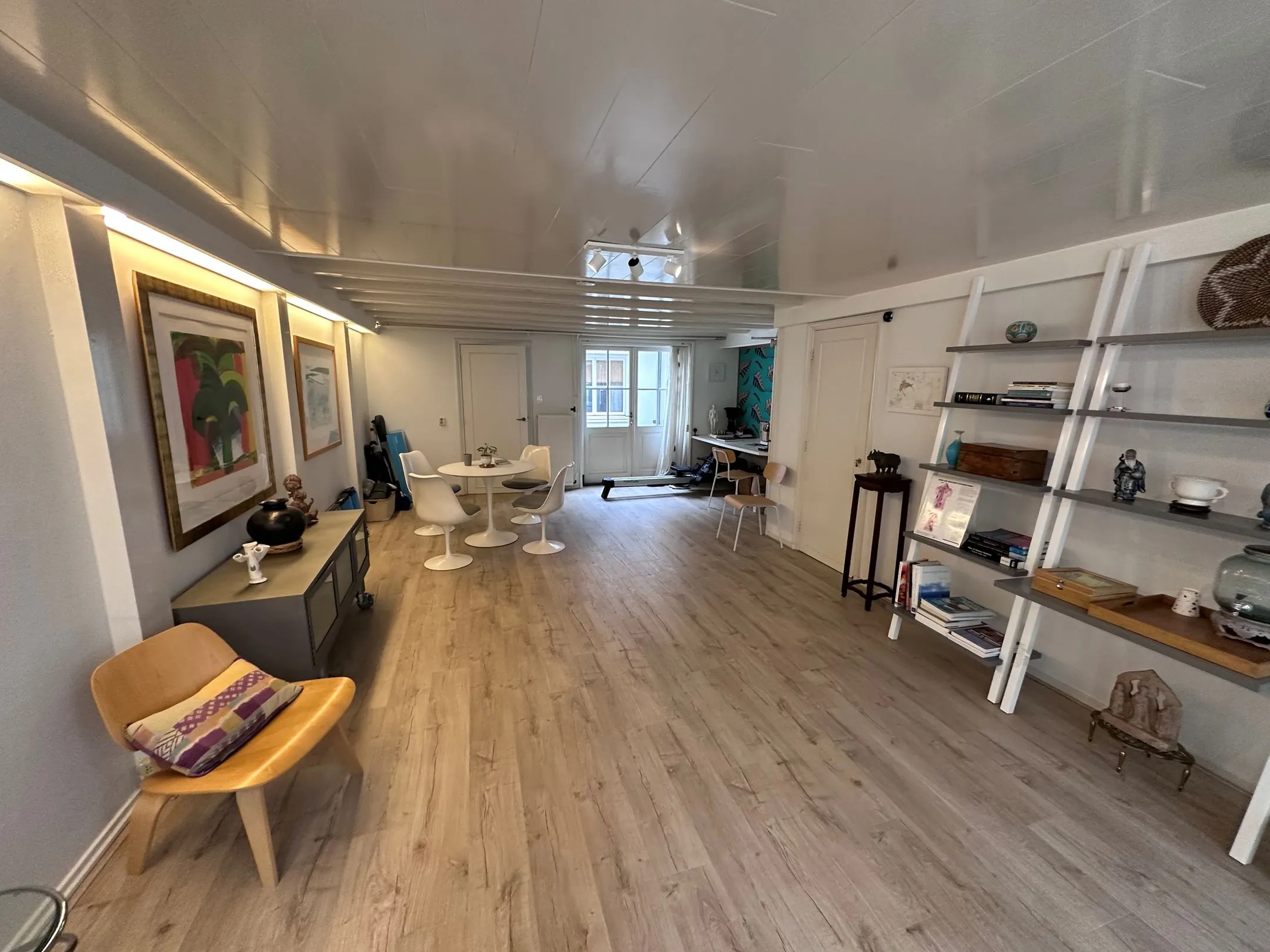 Moderne, langgerekte woonkamer met houten vloer, witte meubels en een open boekenkast tegen de muur.