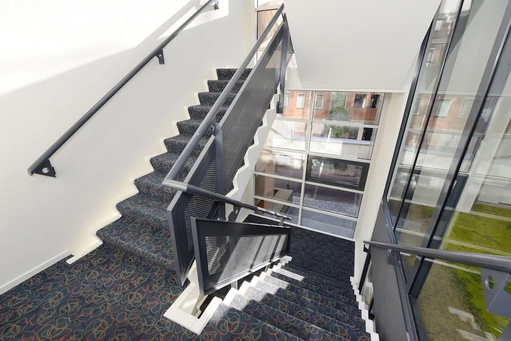 Trapportaal met tapijt en zwarte leuningen in een modern gebouw aan het Spui, met grote ramen en uitzicht op de straat.