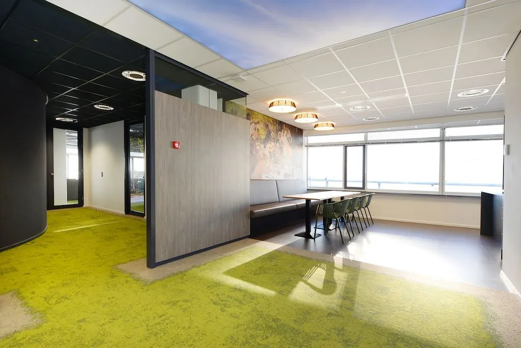 Modern kantoorinterieur met groene vloerbedekking, zithoek met lange tafel en wanddecoratie nabij raam met veel daglicht.