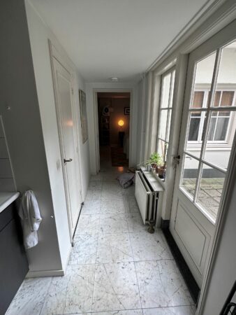 Een smalle, lichte gang met marmeren vloer, een open deur naar een kamer met een brandende lamp en een radiator onder het raam.