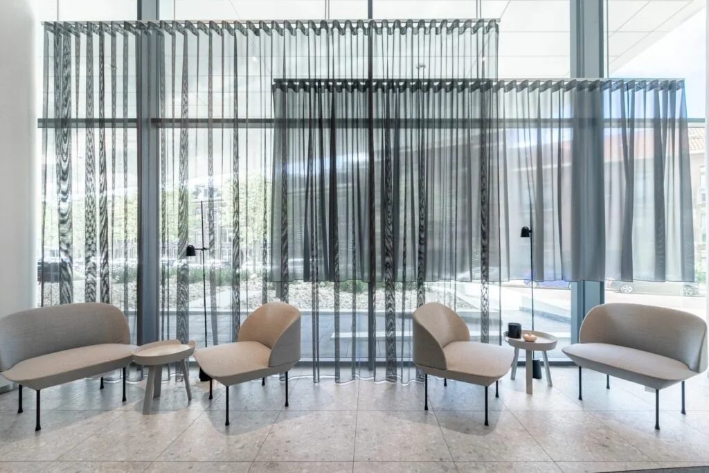 Moderne wachtruimte met beige zitmeubels, kleine bijzettafels en transparante grijze gordijnen voor een glazen gevel.