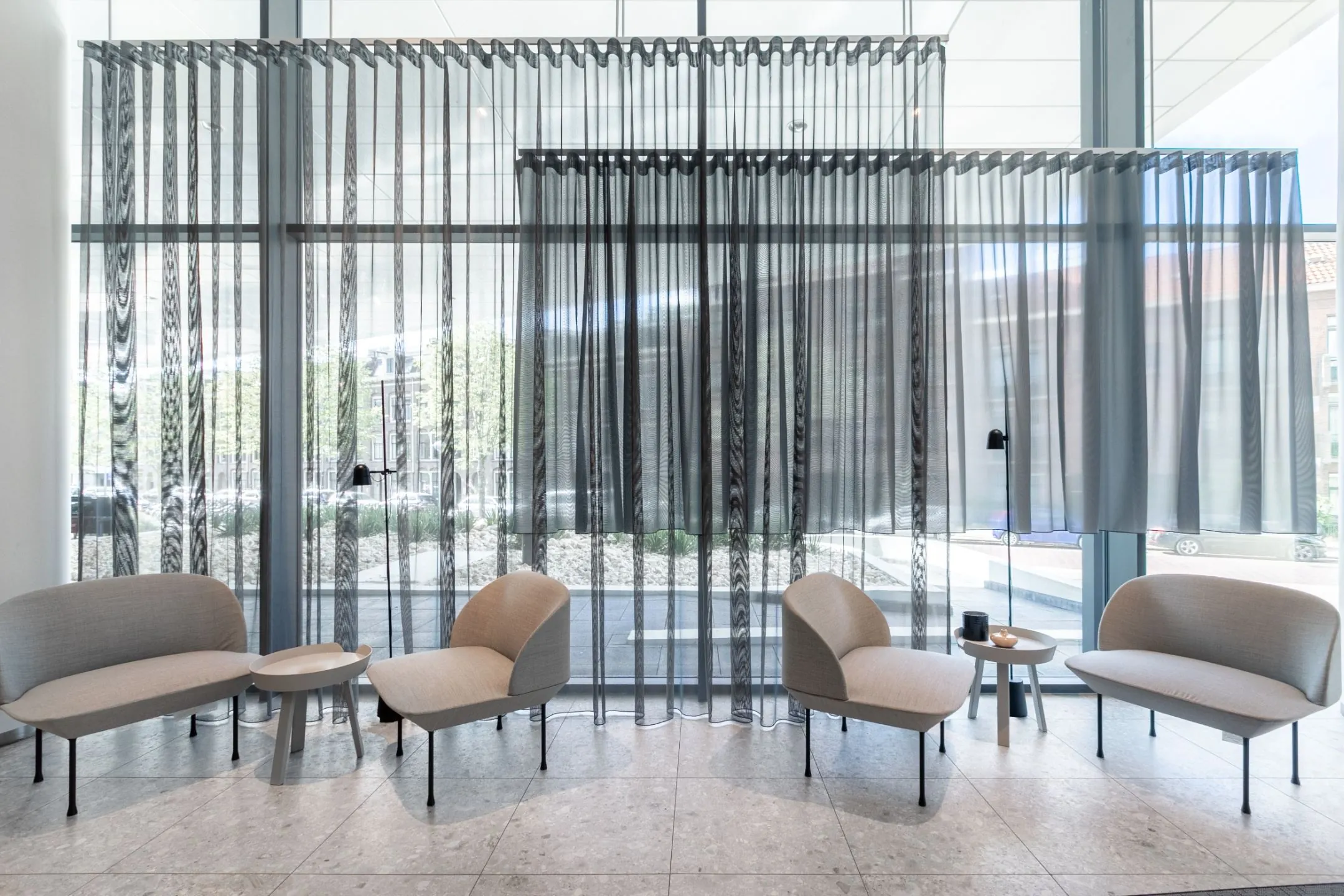 Moderne wachtruimte met beige zitmeubels, kleine bijzettafels en transparante grijze gordijnen voor een glazen gevel.