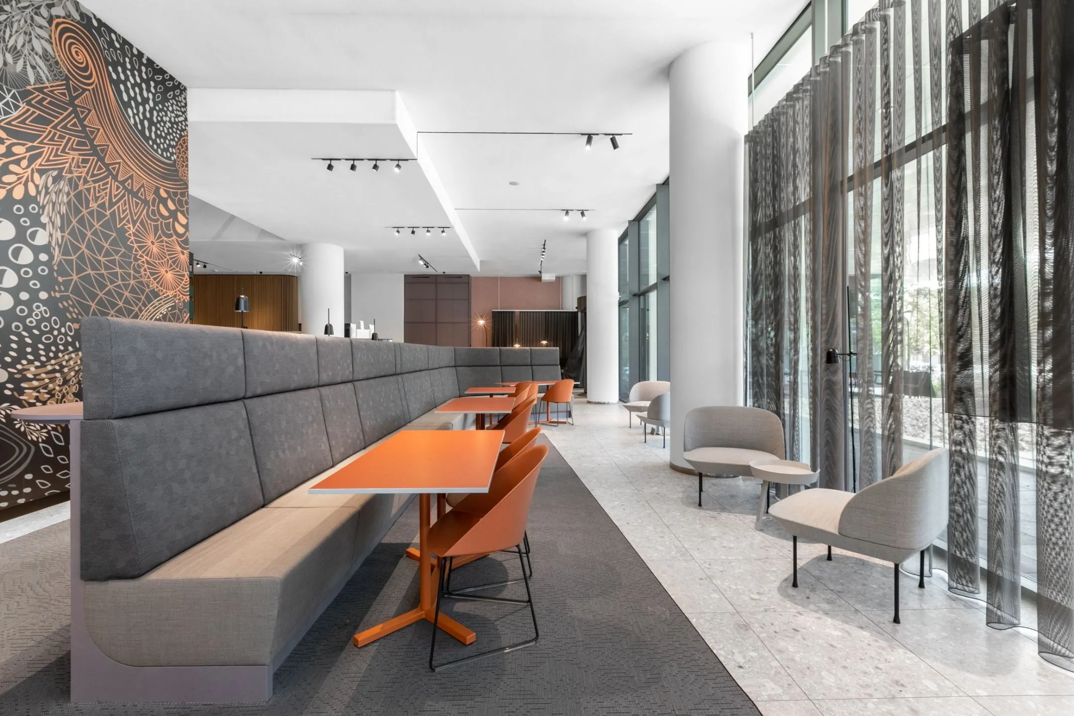 Moderne lounge- en werkruimte met oranje tafels, grijze banken en grote raampartijen met transparante gordijnen.