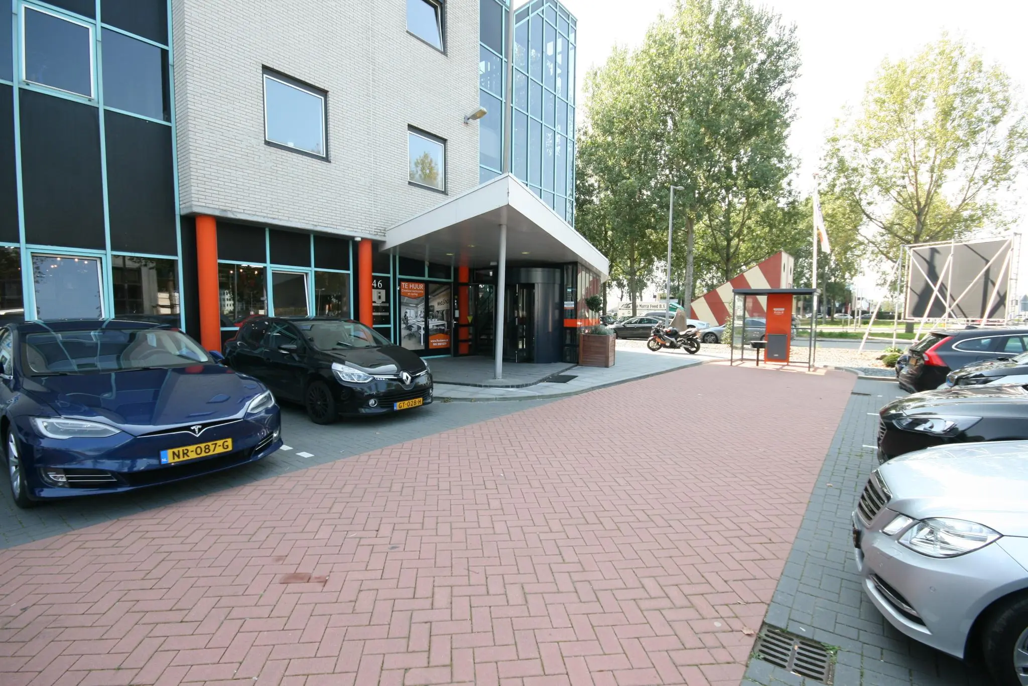Ingang van een kantoorpand aan de Corkstraat met geparkeerde auto’s en een motor bij een bushalte.