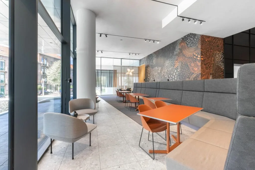 Moderne, lichte lounge- en werkruimte met oranje tafels, grijze banken en een artistieke muurdecoratie.
