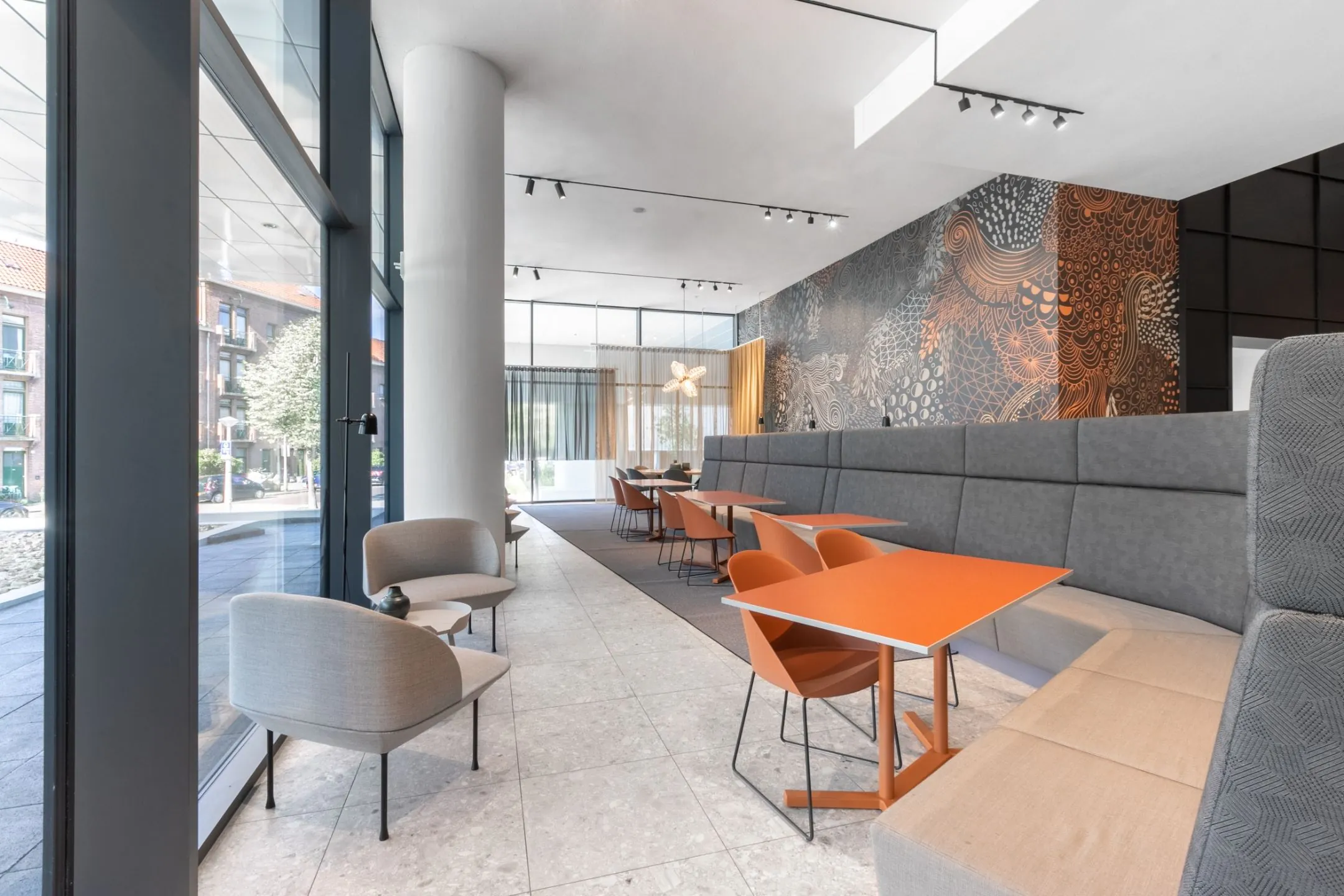 Moderne, lichte lounge- en werkruimte met oranje tafels, grijze banken en een artistieke muurdecoratie.
