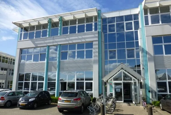Modern kantoorgebouw aan de Gyroscoopweg 144 met glasgevel, geparkeerde auto's en fietsenrekken bij de ingang.