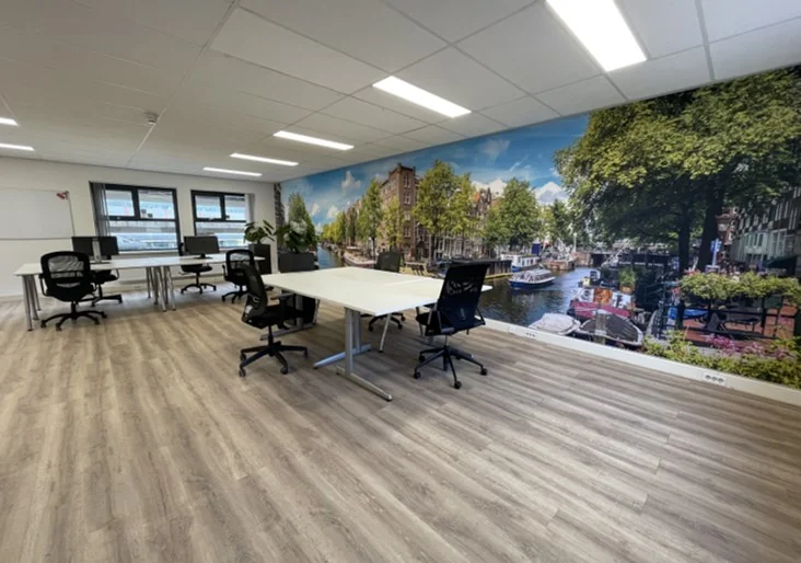 Moderne kantoorruimte aan de Arlandaweg met werkplekken, laminaatvloer en een muurfoto van een Amsterdams grachtenbeeld.