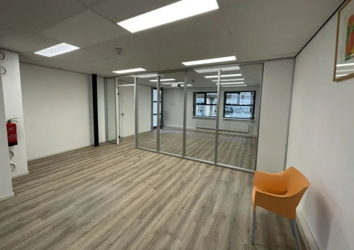 Lichte, lege kantoorruimte aan de Arlandaweg met glazen tussenwand, houten vloer en een oranje stoel.