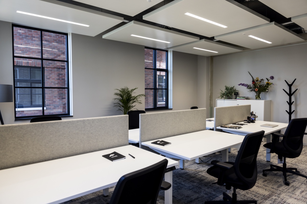 Moderne kantoorruimte met bureaus, stoelen en planten naast grote ramen en een grijze wand.
