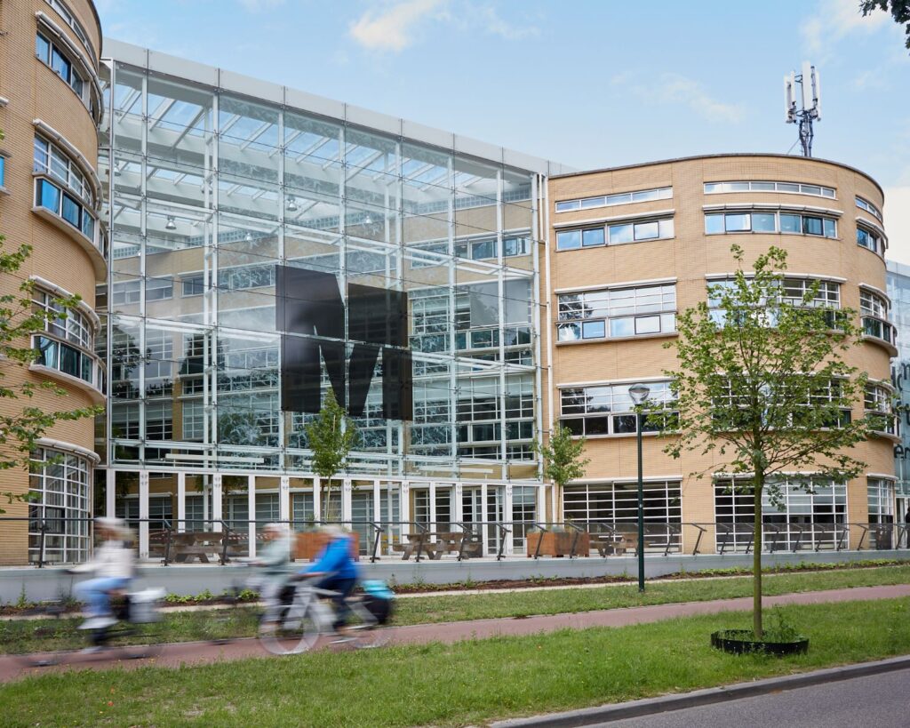 Moderne glazen gevel van het AKN-gebouw in Hilversum met voorbijfietsende mensen op de voorgrond.