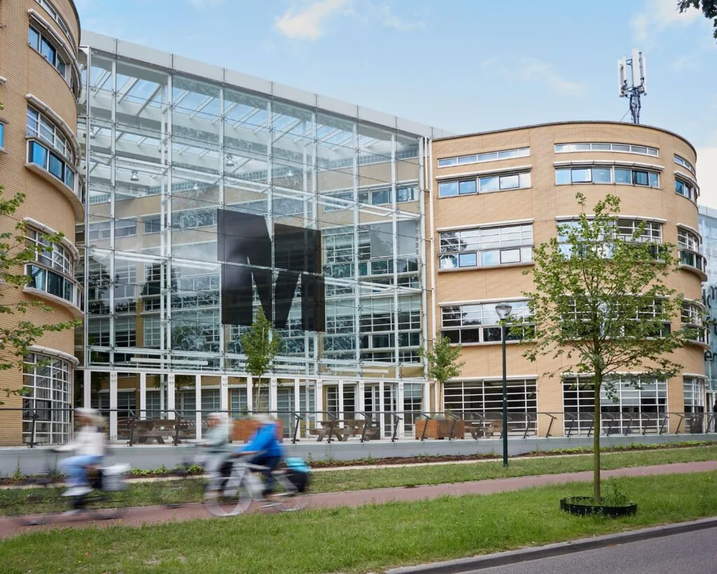 Moderne glazen gevel van het AKN-gebouw in Hilversum met voorbijfietsende mensen op de voorgrond.