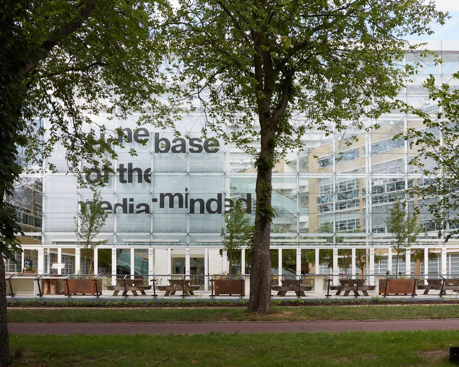 Glazen gevel van het AKN-gebouw in Hilversum met grote tekst “the base for the media-minded” en picknickbanken op het terras ervoor.